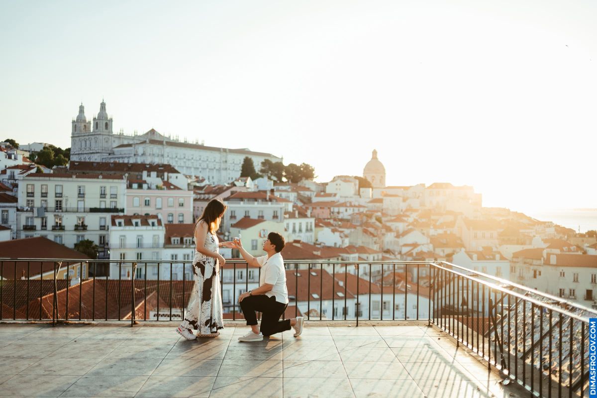 Lisbon proposal! Couple on a balcony with Alfama rooftops. Man proposes to woman with Lisbon cityscape background. Romance in Portugal.
