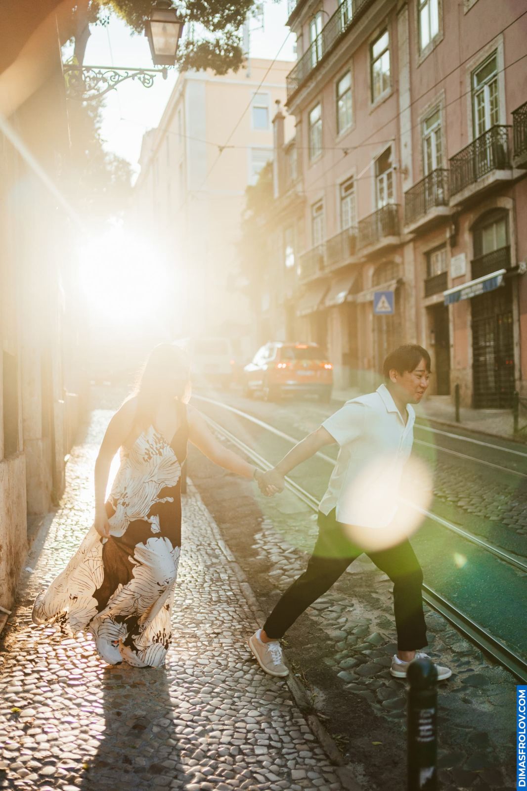 Couple holding hands crossing tram tracks in sunny Lisbon. Cobblestone street, historic buildings. Romantic travel.