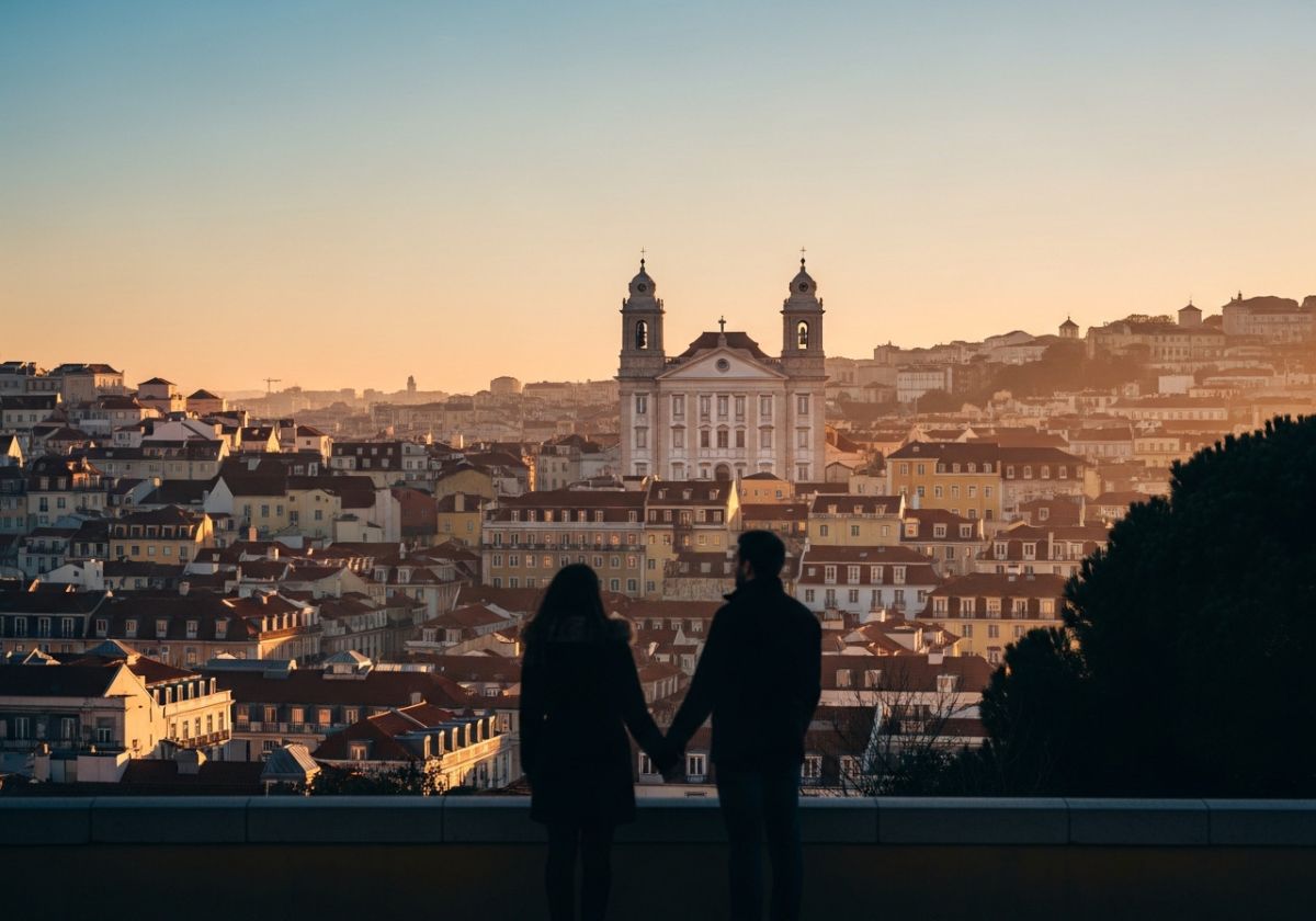 A wide shot of Lisbon during winter sunset, with warm colors in the sky and soft light reflecting on the buildings. A couple is silhouetted in the foreground, holding hands and looking at the view. Focus on the light and the city-s atmosphere.