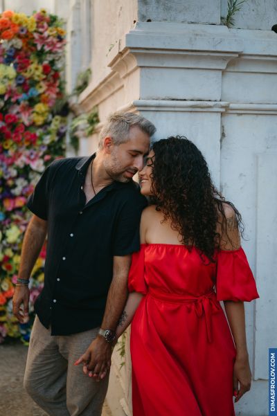 Couple in Lisbon. Woman in red dress leans against a white wall adorned with flowers. Romantic moment in Portugal.