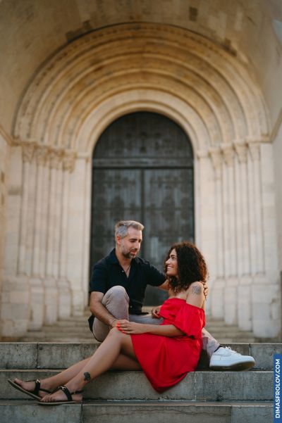 Couple in Lisbon: Woman in red dress sits with man on stone steps of a historic building with arched doorway. Romantic moment.