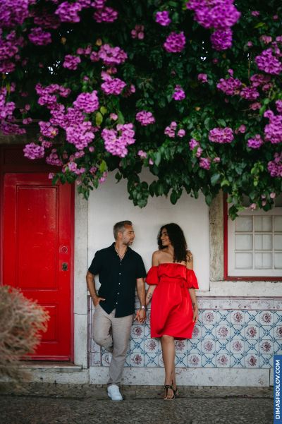 Couple in Lisbon. Woman in red dress, man in black shirt, standing by tiled wall with purple flowers. Romantic Portugal scene.