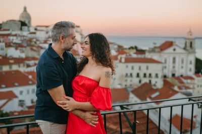 Couple in Lisbon, Portugal. Woman in red dress, man embracing her. Alfama rooftops & Tagus River in background. Romantic Lisbon scene.