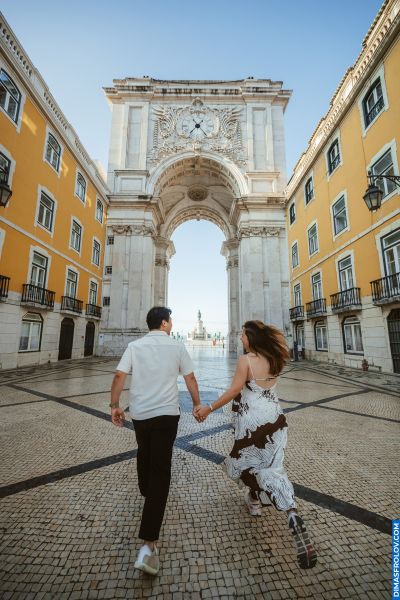 Couple running hand-in-hand towards Arco da Rua Augusta in Lisbon, Portugal. Cobblestone streets, yellow buildings, romantic travel.