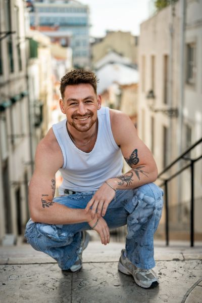 Man with tattoos squats on Lisbon steps. Alfama street scene in background. Portugal travel.