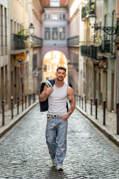Man walks on cobblestone street in Lisbon, Portugal. European architecture, jacket over shoulder, tattoos.