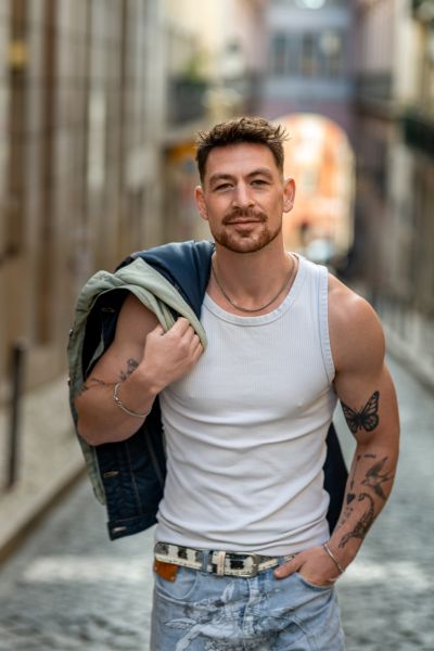Man in Lisbon alley. White tank top, jacket over shoulder, tattoos. Cobblestone street, European vibe.