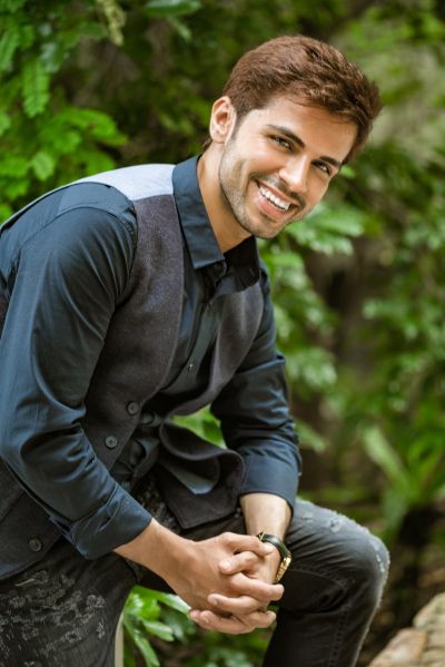 Smiling man in Lisbon garden. Dark shirt, vest, and jeans. Lush green background. Portugal travel. Men-s fashion.
