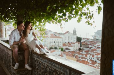 Couple enjoys Lisbon view from tiled terrace. Alfama rooftops & church in background. Romantic travel in Portugal.