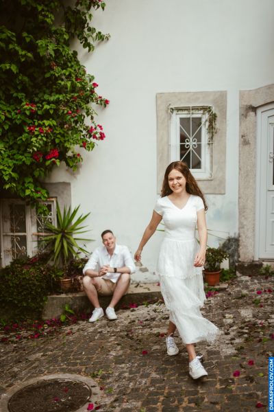Couple in Lisbon. Woman in white dress walks on cobblestone street, man sits on steps. Alfama vibes, bougainvillea, Portugal.